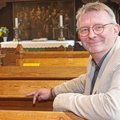 Ein Mann mit Brille in hellem Anzug sitzt in einer Kirchenbank und lächelt in die Kamera
